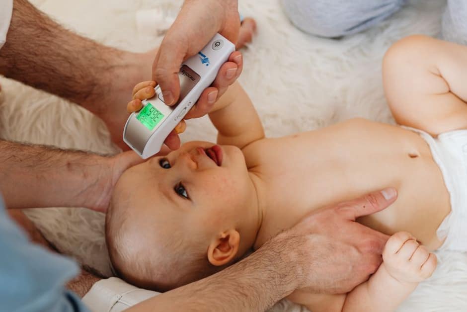 Parent posant la main sur le front de son bébé allongé dans son lit, thermomètre posé à proximité