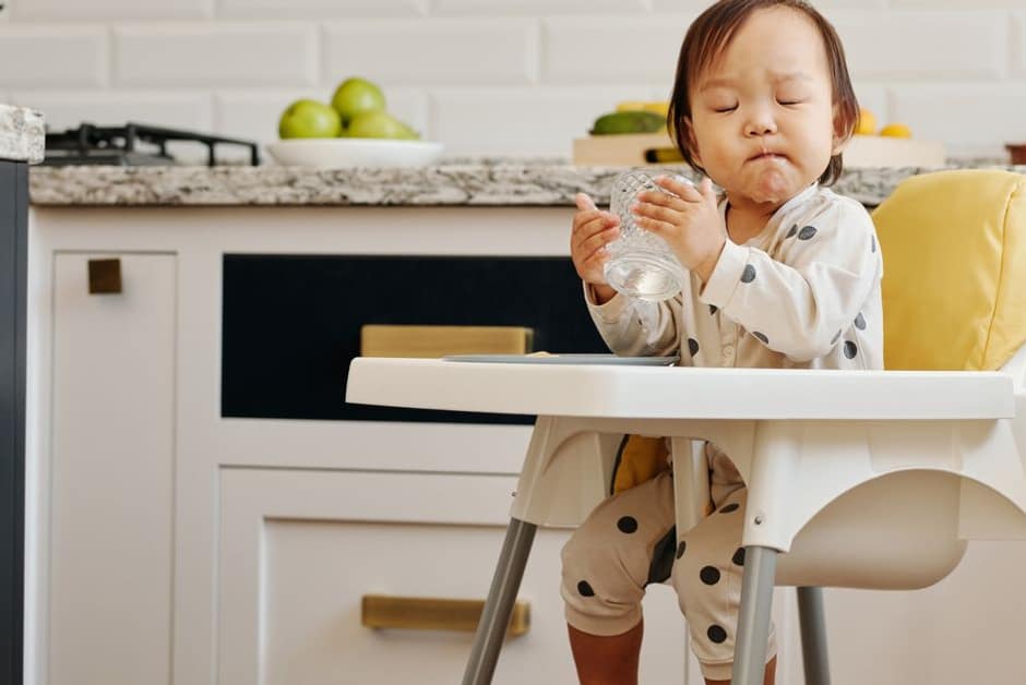 Tout-petit buvant un verre d'eau dans sa chaise haute : l'hydratation, priorité de la gastro