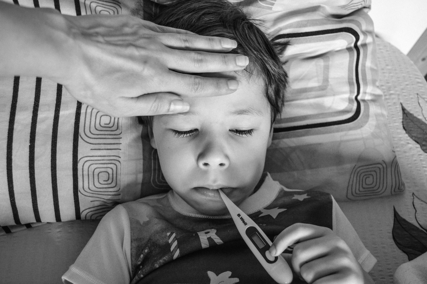 Parent posant la main sur le front d'un jeune enfant fiévreux allongé, thermomètre à la bouche