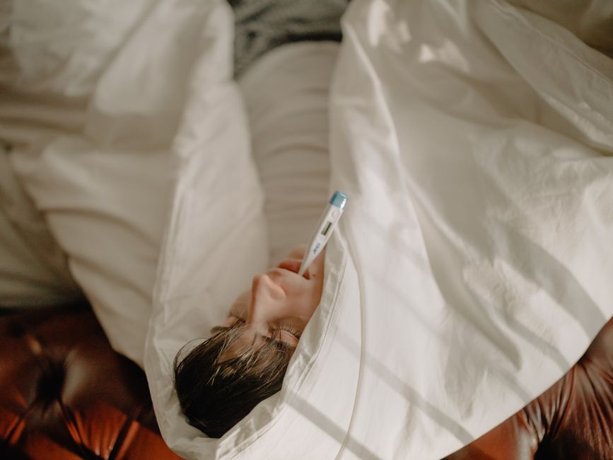 Adulte allongé sous une couette, un thermomètre à la bouche, en train de surveiller sa température à la maison